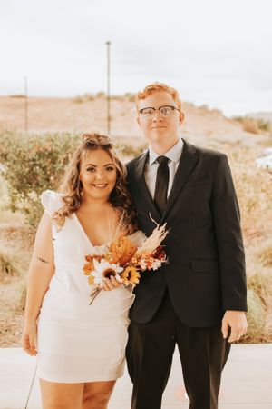 Newlyweds pose together in formal attire with autumn-colored bouquet against desert backdrop.