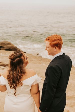 A couple in wedding attire stands together on a rocky beach overlooking the ocean.