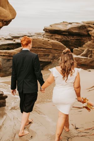 A couple walks barefoot along rocky coastal cliffs holding hands in wedding attire.