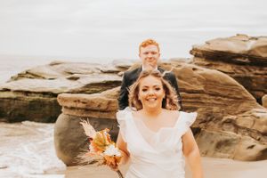 A joyful wedding moment captured on rocky beach terrain with fall-colored bouquet.