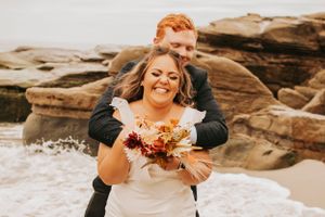 A playful wedding photo showing laughter and embraces on a rocky beach landscape.