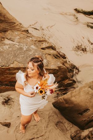 An overhead shot of bride in white dress holding sunflowers on rocky beach terrain.