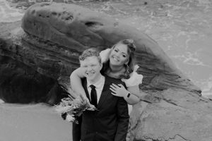 A black and white wedding portrait on rocky coastline with ocean waves in background.