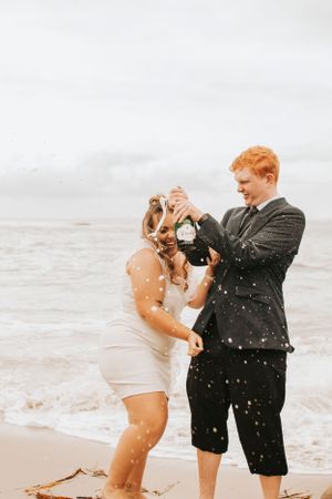 Joyful couple celebrating with champagne on a misty beach during golden hour.