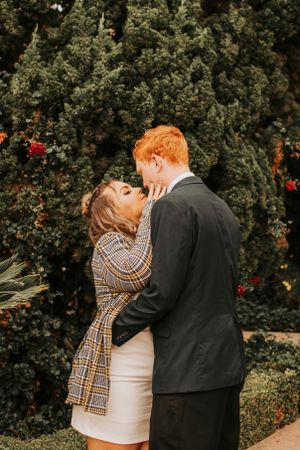 Romantic couple embracing in front of lush green bushes with red flowers.