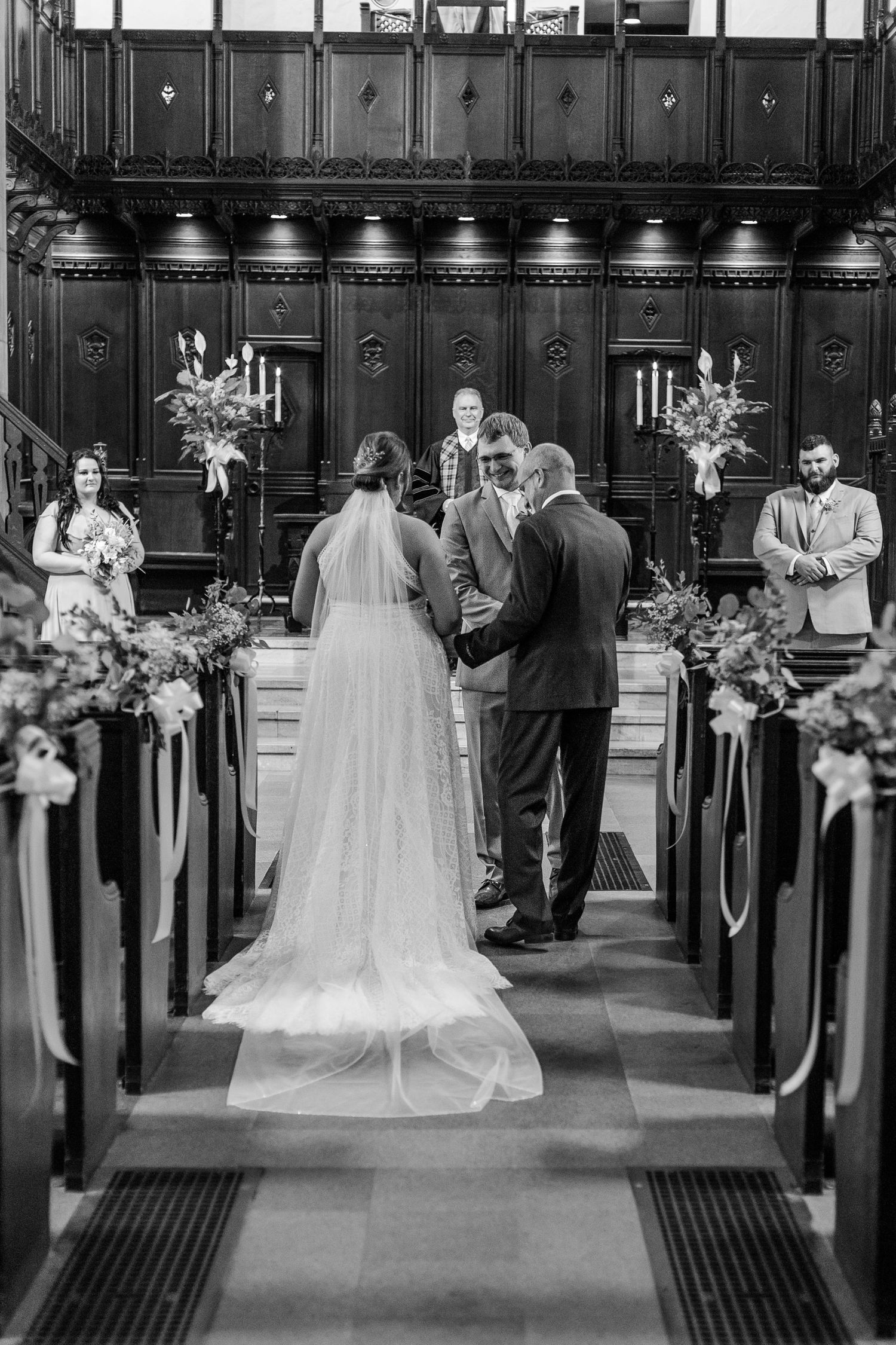 Wedding ceremony taking place in a church aisle with dark wooden pews decorated with floral arrangements.