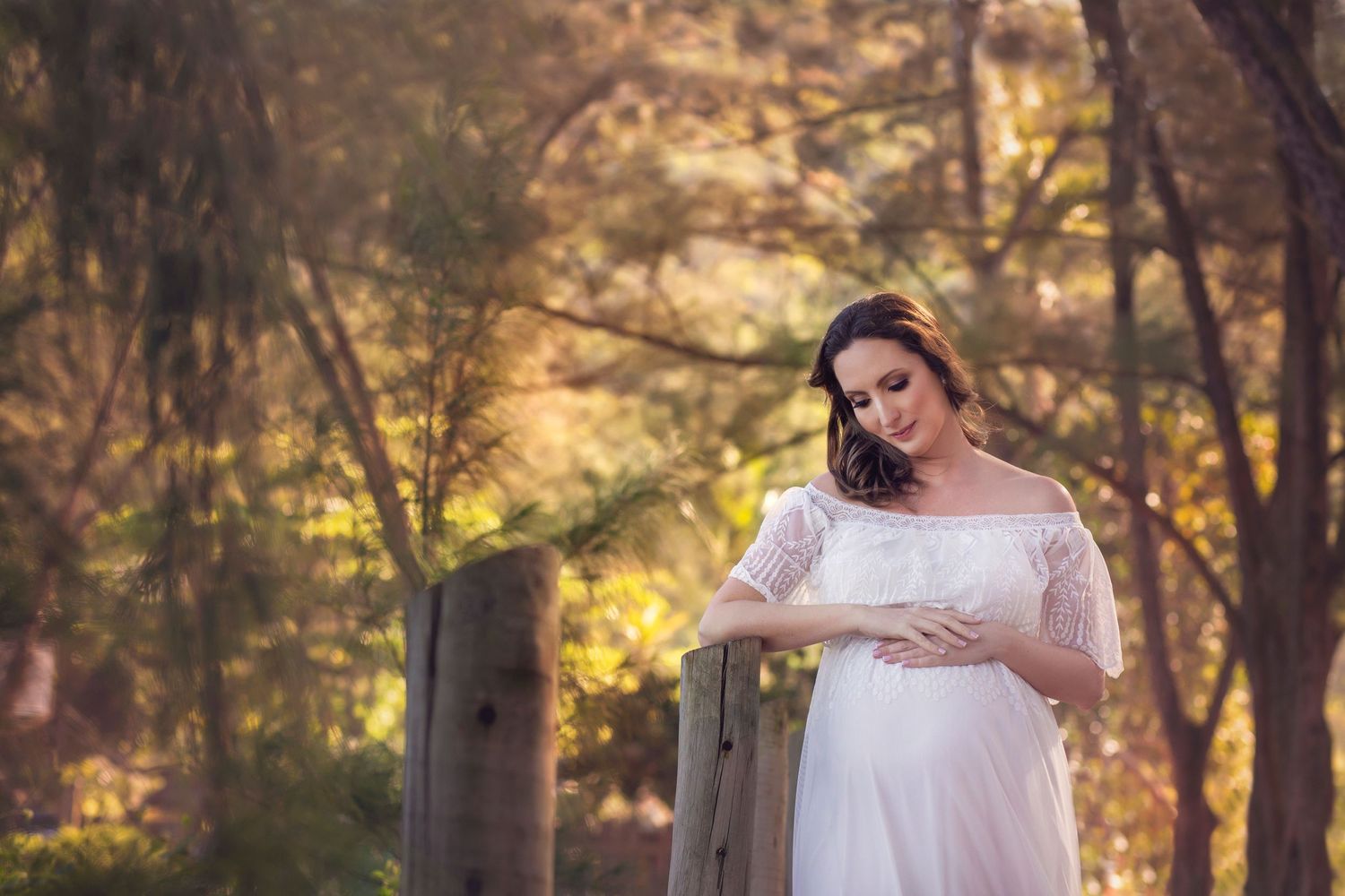Maternity portrait of woman in white dress glowing in sunlit forest setting.
