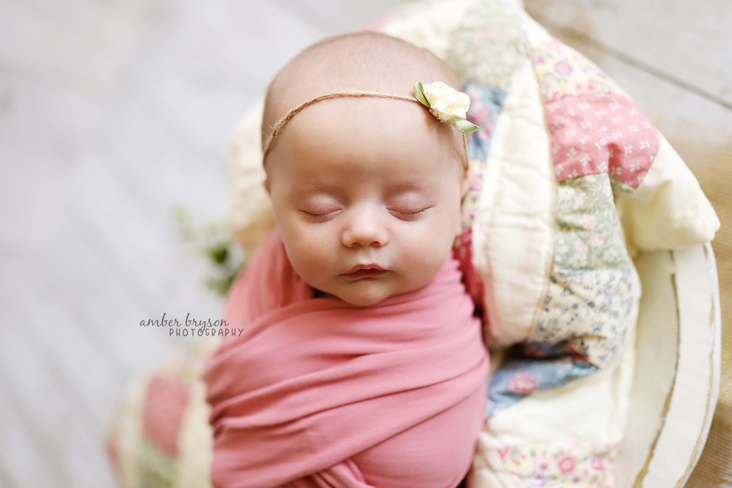 Sleeping newborn in pink wrap with floral quilted backdrop.