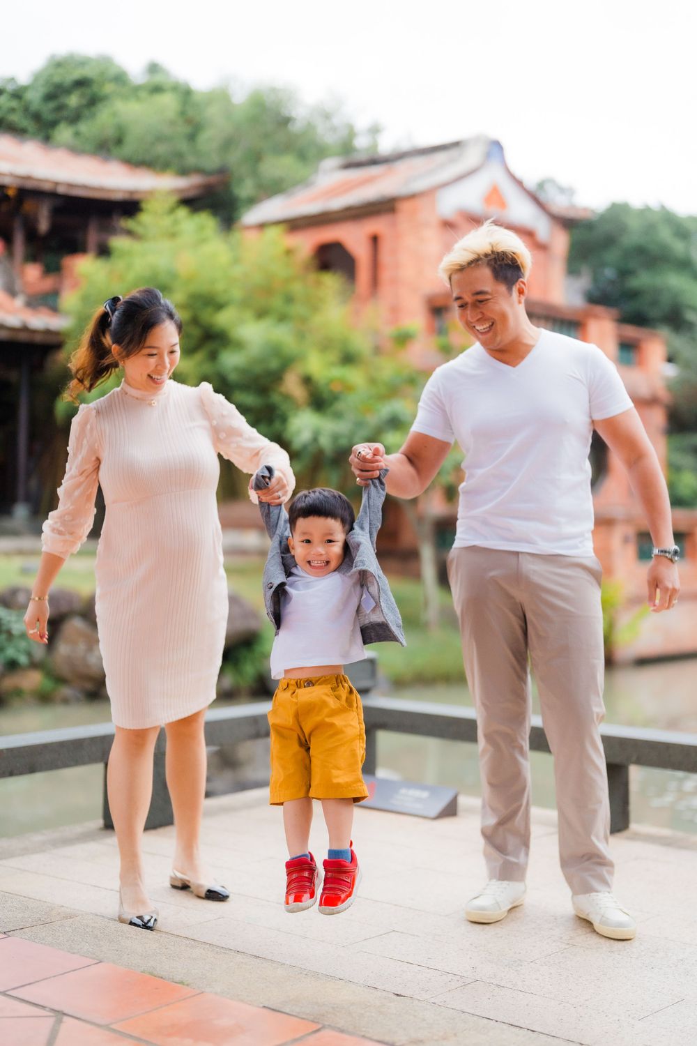 Family helps young child jump while walking along outdoor path near traditional buildings.
