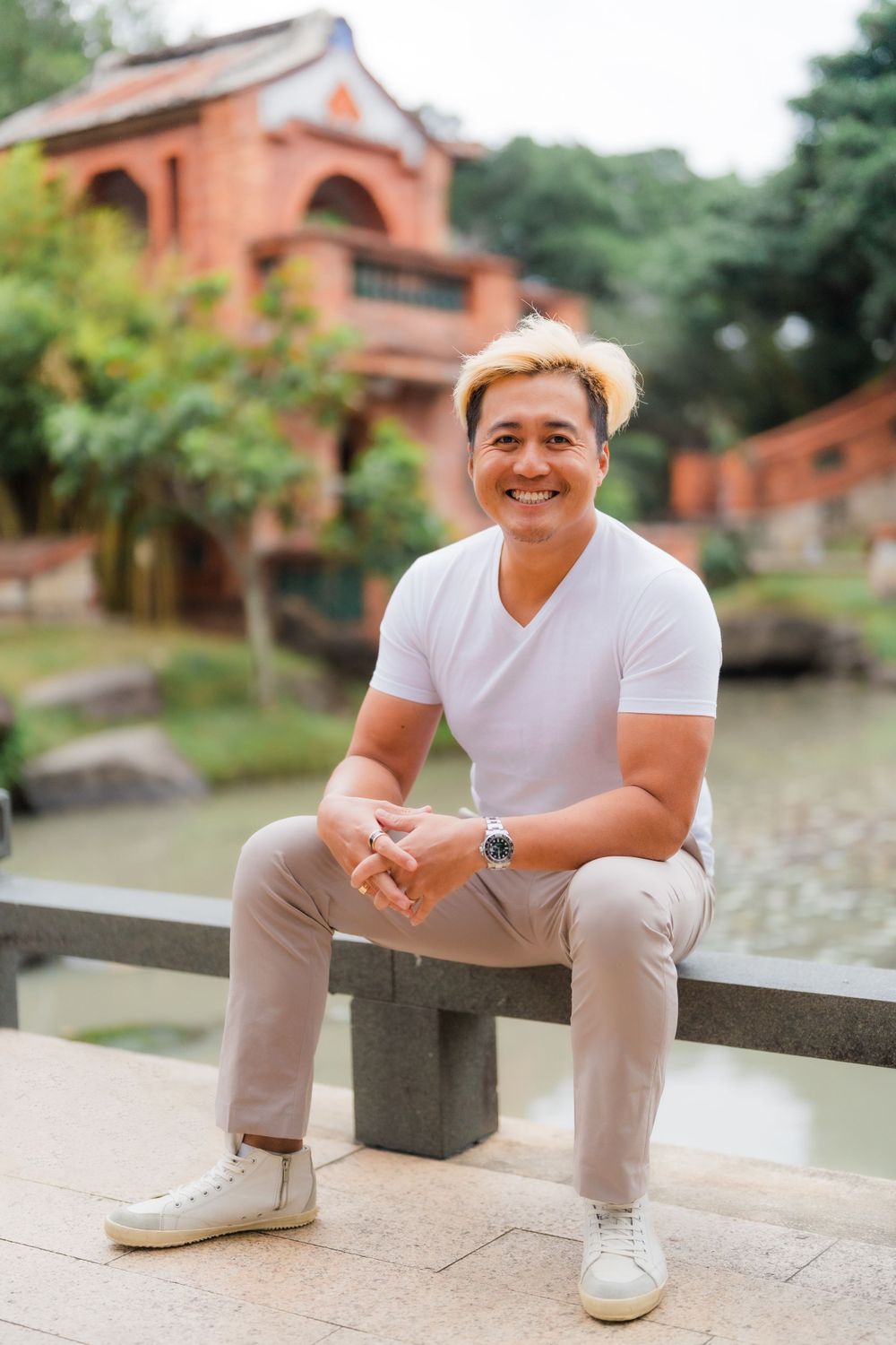 Man in white t-shirt and khaki pants sitting on stone bench with Asian architecture behind.