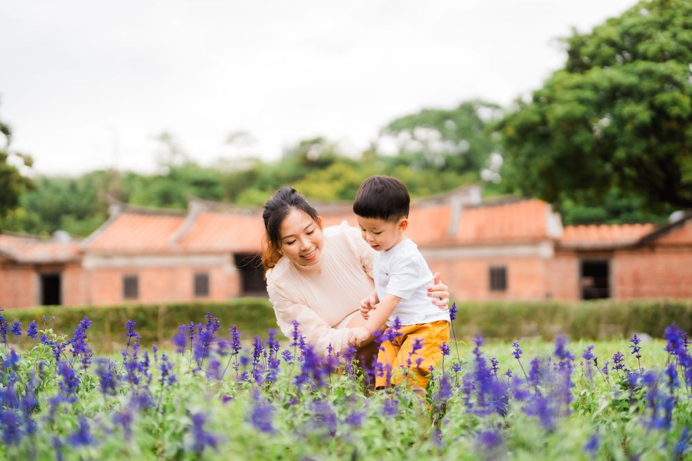 Parent and child enjoying purple wildflowers with traditional brick buildings in background.