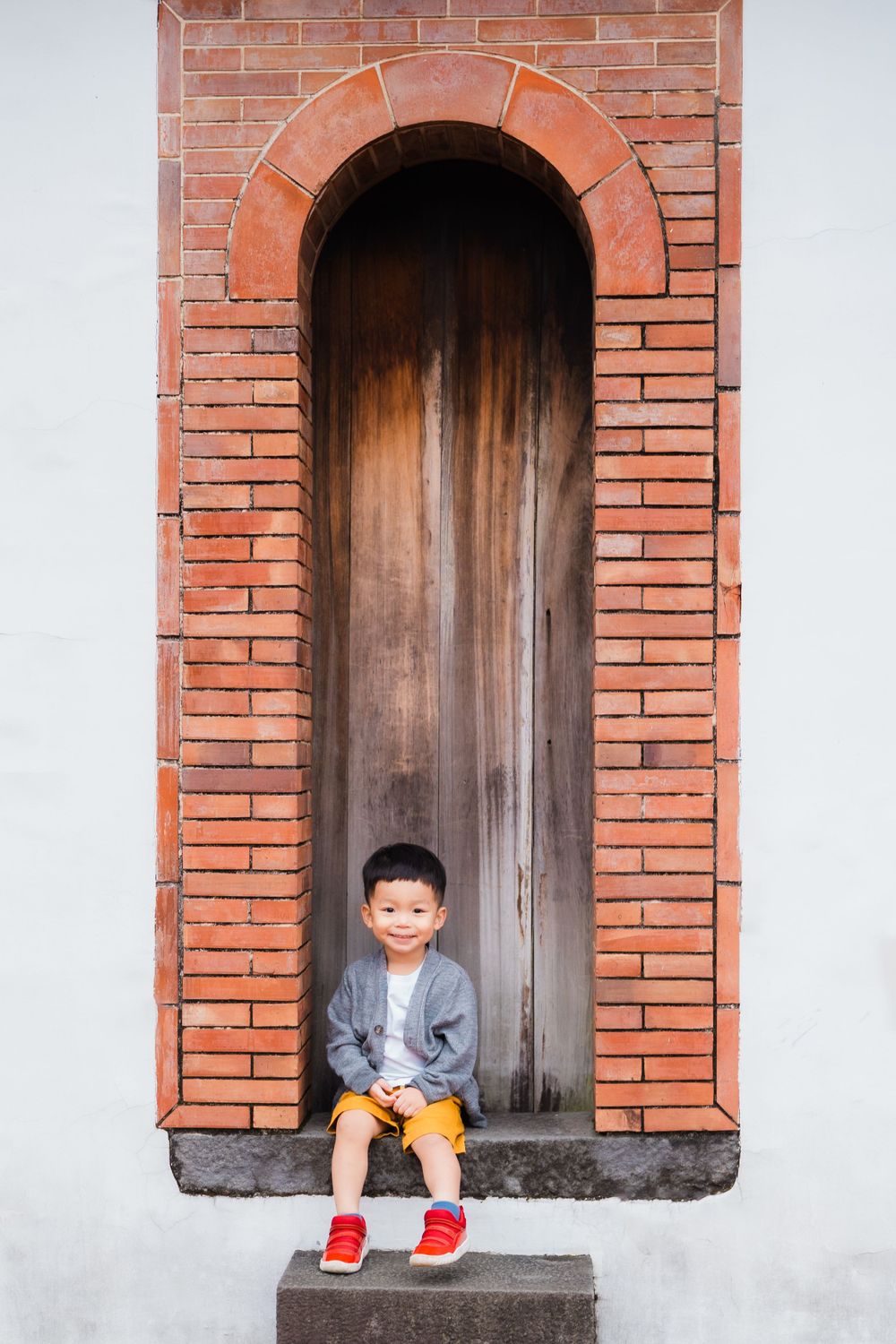 Child in gray jacket and red shoes sits in brick archway doorframe.