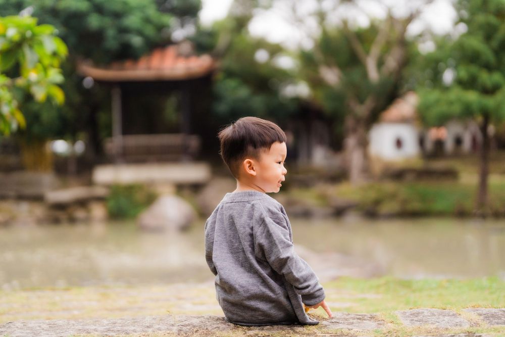 Young child in gray sweater sitting alone by a pond with traditional buildings in background.