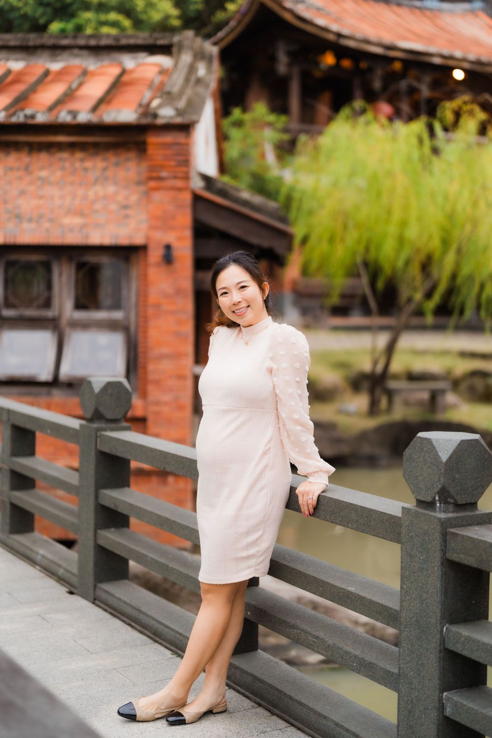 Woman in cream dress posing on wooden bridge with traditional Asian architecture in background.