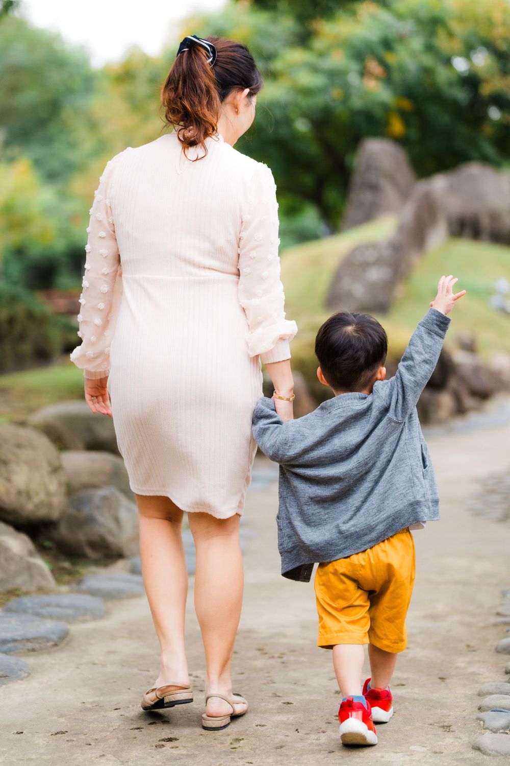Mother and child walking on a garden path, with the child wearing yellow shorts and a gray sweater reaching up.