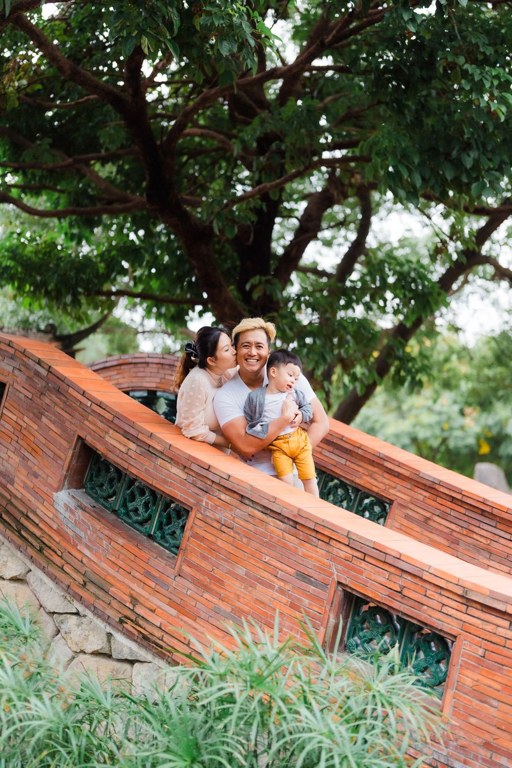 Family sitting together on a rustic orange brick bridge under a large tree.