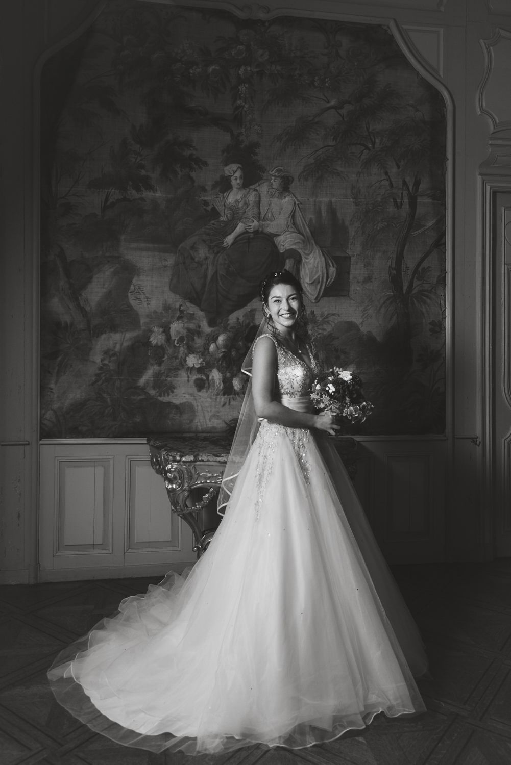 Black and white portrait of bride in flowing gown standing before ornate wall mural.