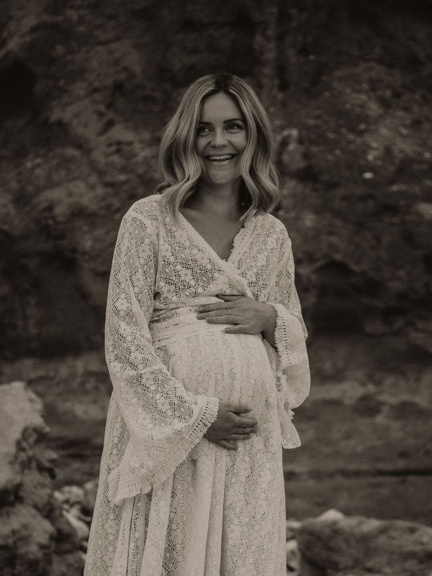 A dreamy black and white maternity portrait showcases a flowing lace dress against rugged cliff walls.