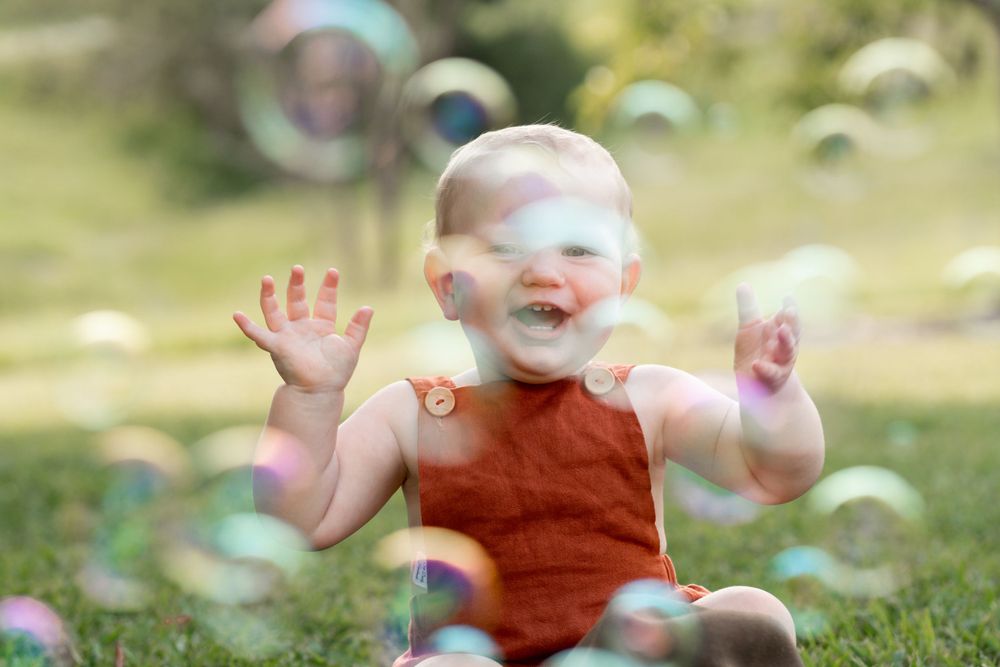 Baby sits laughing on the grass, surrounded by a cloud of bubbles