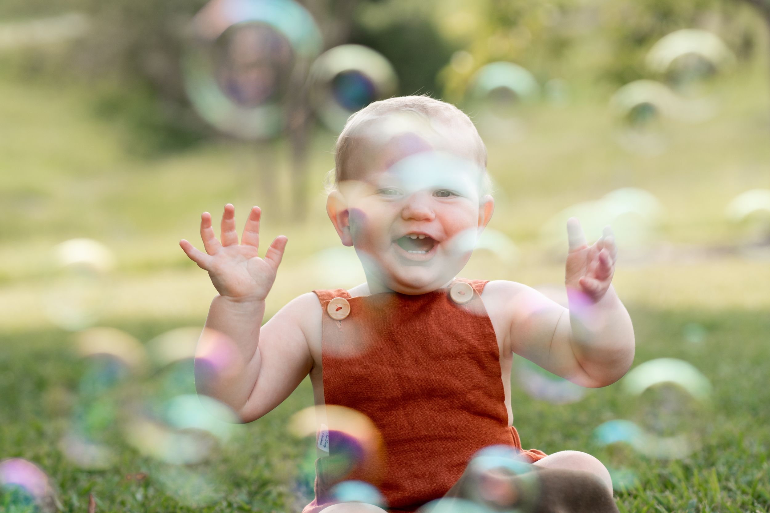 Baby sits laughing on the grass, surrounded by a cloud of bubbles