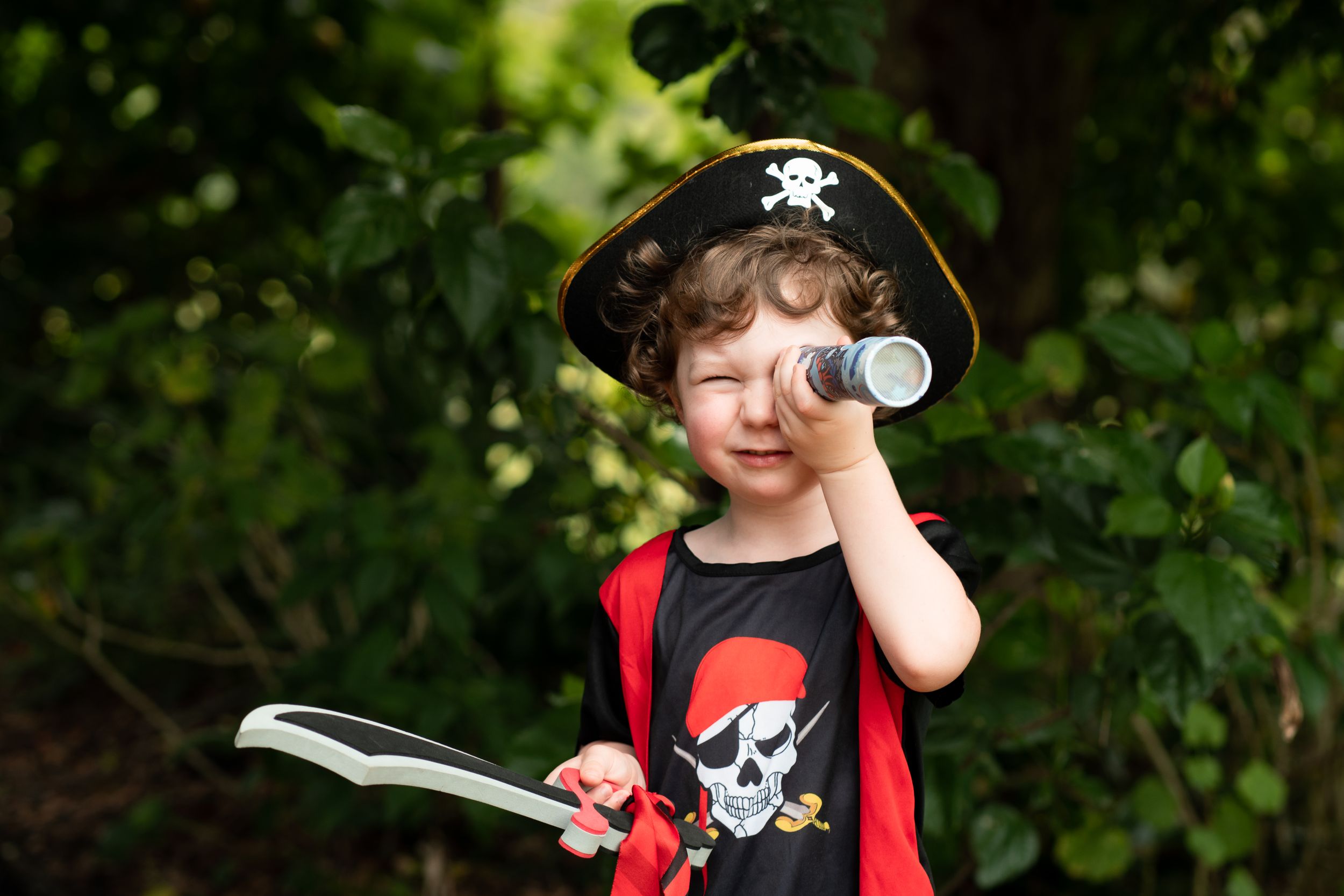 Young boy wearing a pirate costume, peering through a telescope and waving a sword