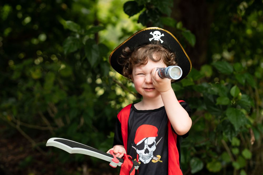 Young boy wearing a pirate costume, peering through a telescope and waving a sword