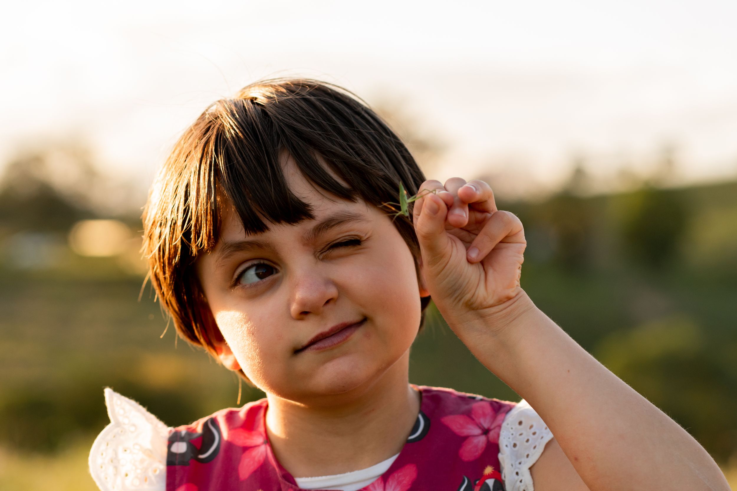 Young girl holds a grasshopper in two fingers at eye level, examining it closely with one eye closed.