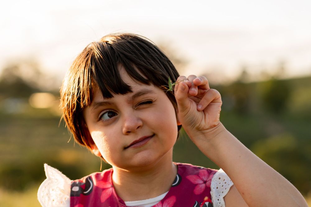 Young girl holds a grasshopper in two fingers at eye level, examining it closely with one eye closed.