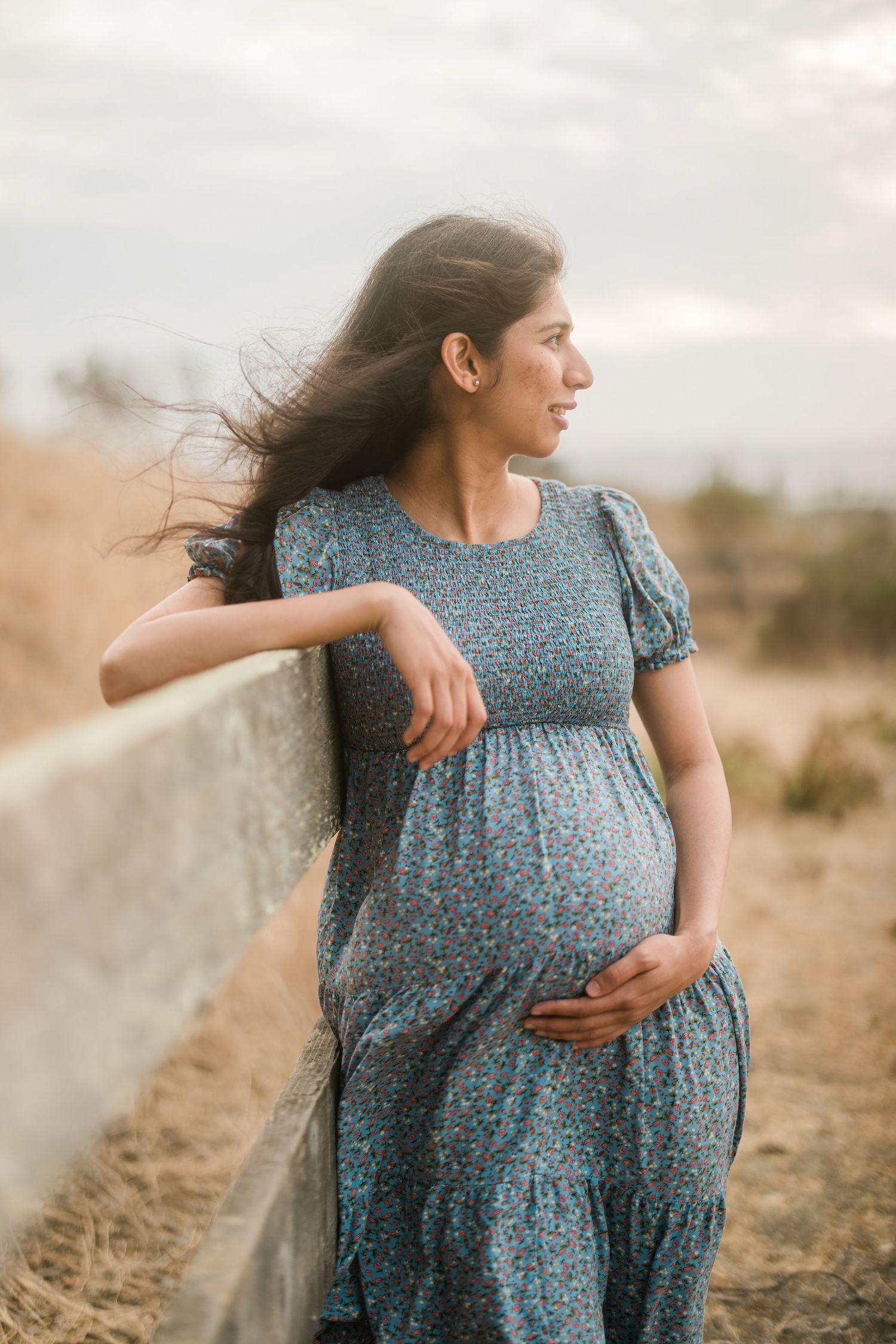 Pregnant woman in blue floral dress leaning against fence during golden hour.