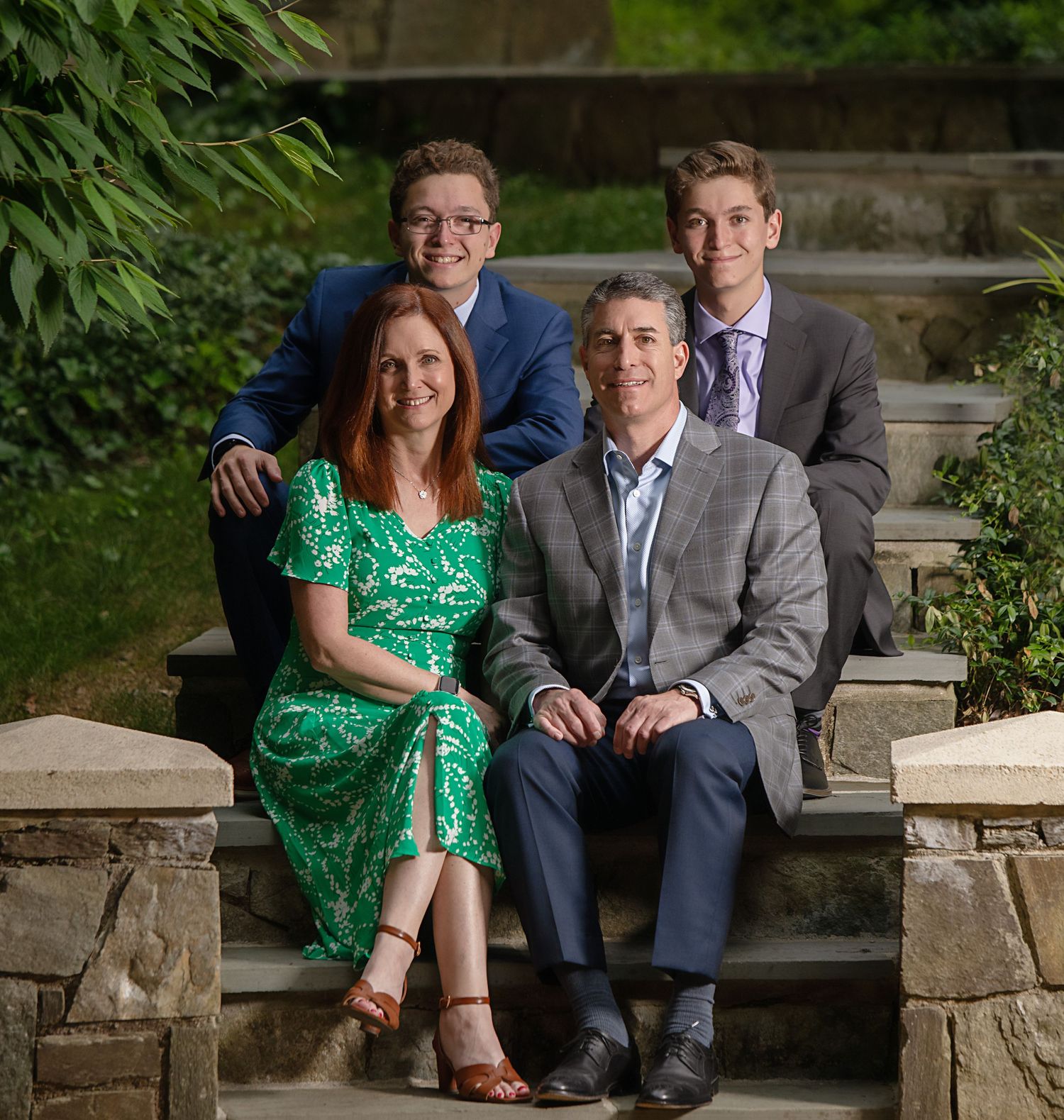 A family group poses together on stone steps outdoors with greenery in the background.
