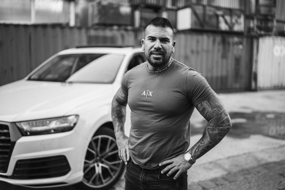 Muscular tattooed person in grey t-shirt standing beside white luxury car.