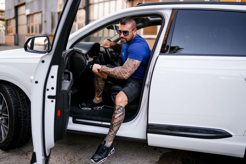 Athletic person with tattoos sitting in doorway of white luxury SUV.