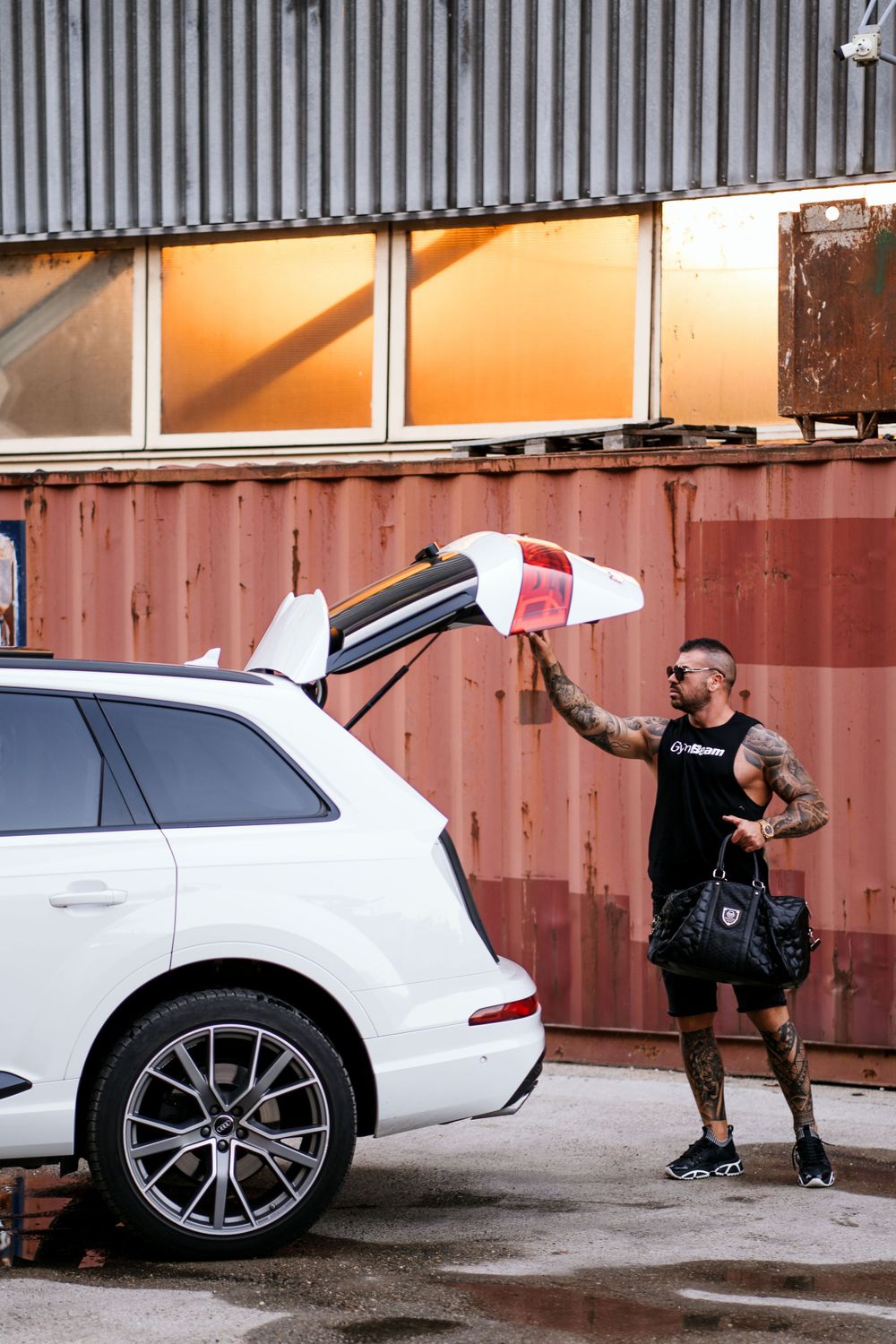 Person in black clothing reaching up to close SUV trunk door near industrial containers.