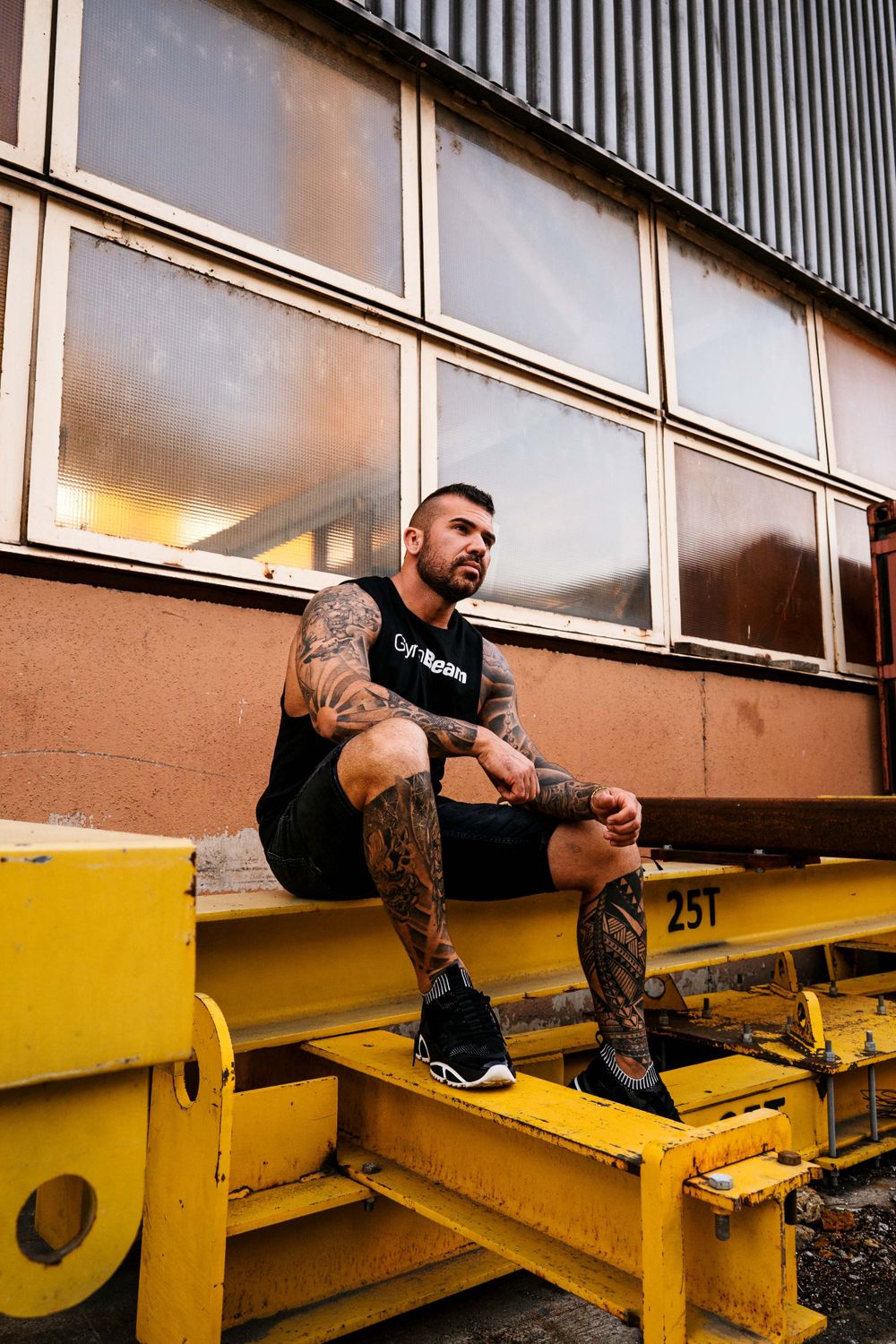 Athletic figure with tattoos sitting on yellow industrial equipment in an urban setting.