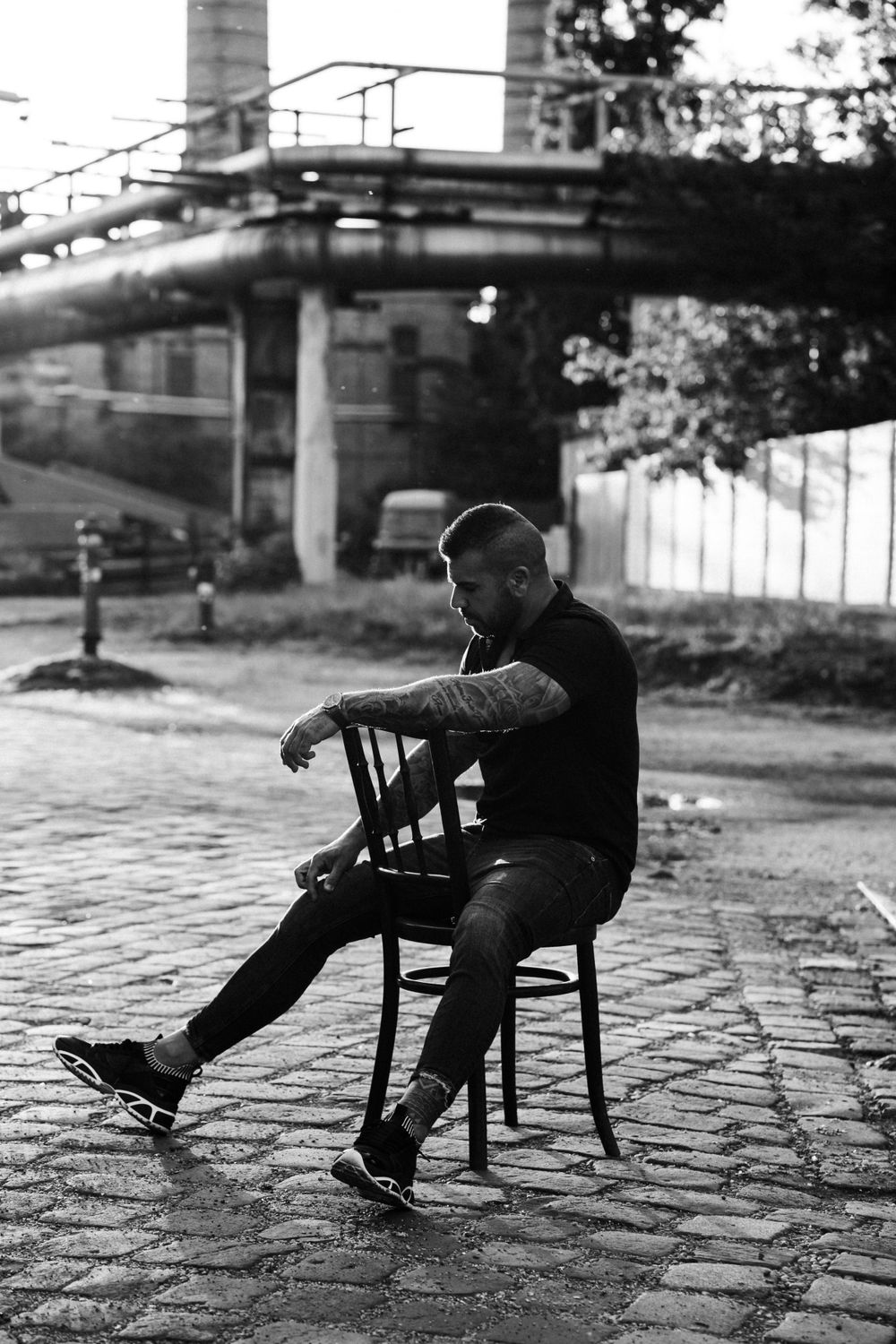 Black and white portrait of person seated on chair against industrial landscape.