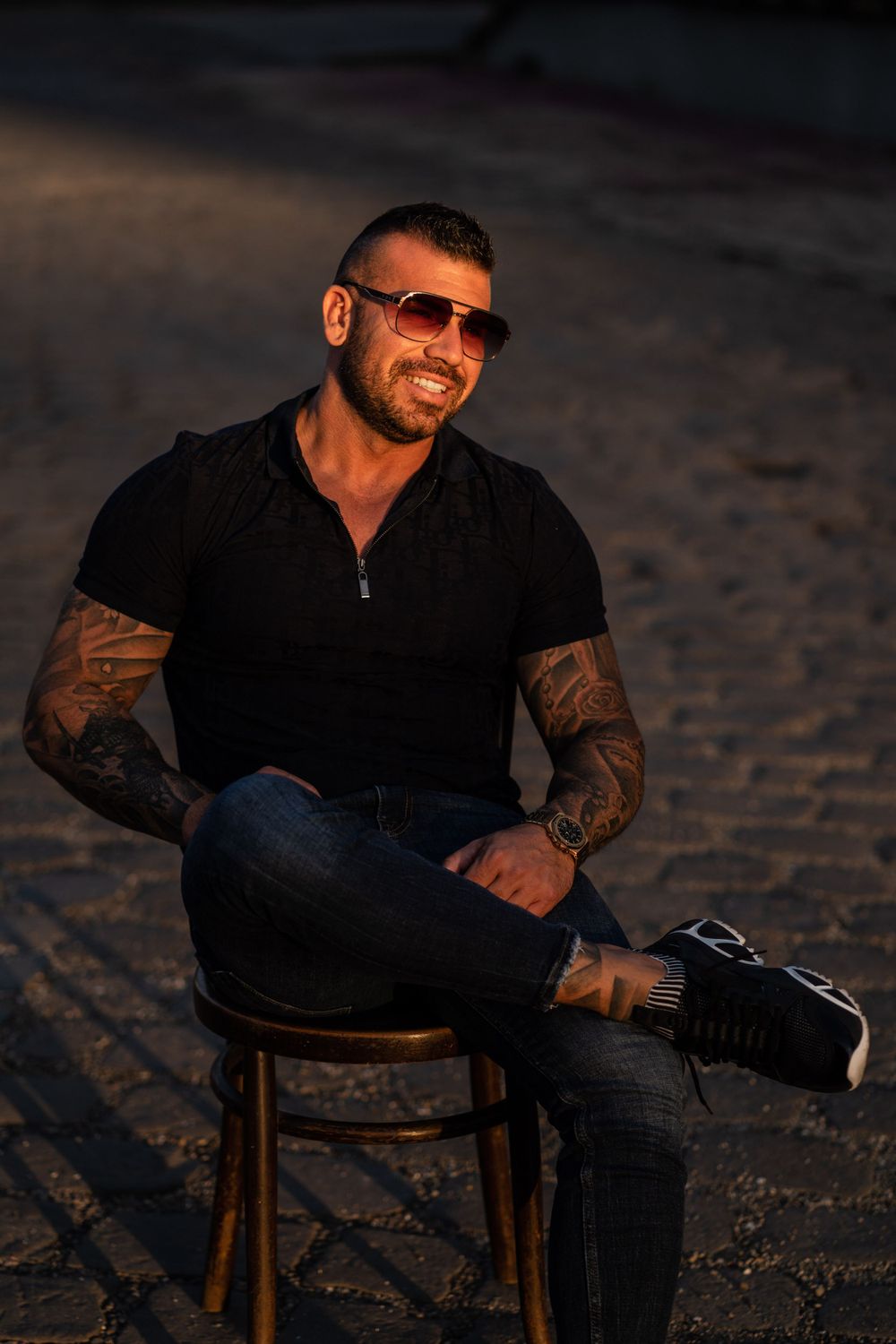 Person in sunglasses and black zip-up jacket seated on wooden chair at dusk.