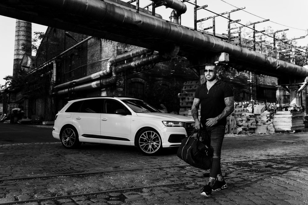 White luxury SUV against industrial backdrop with metal pipelines and urban architecture.