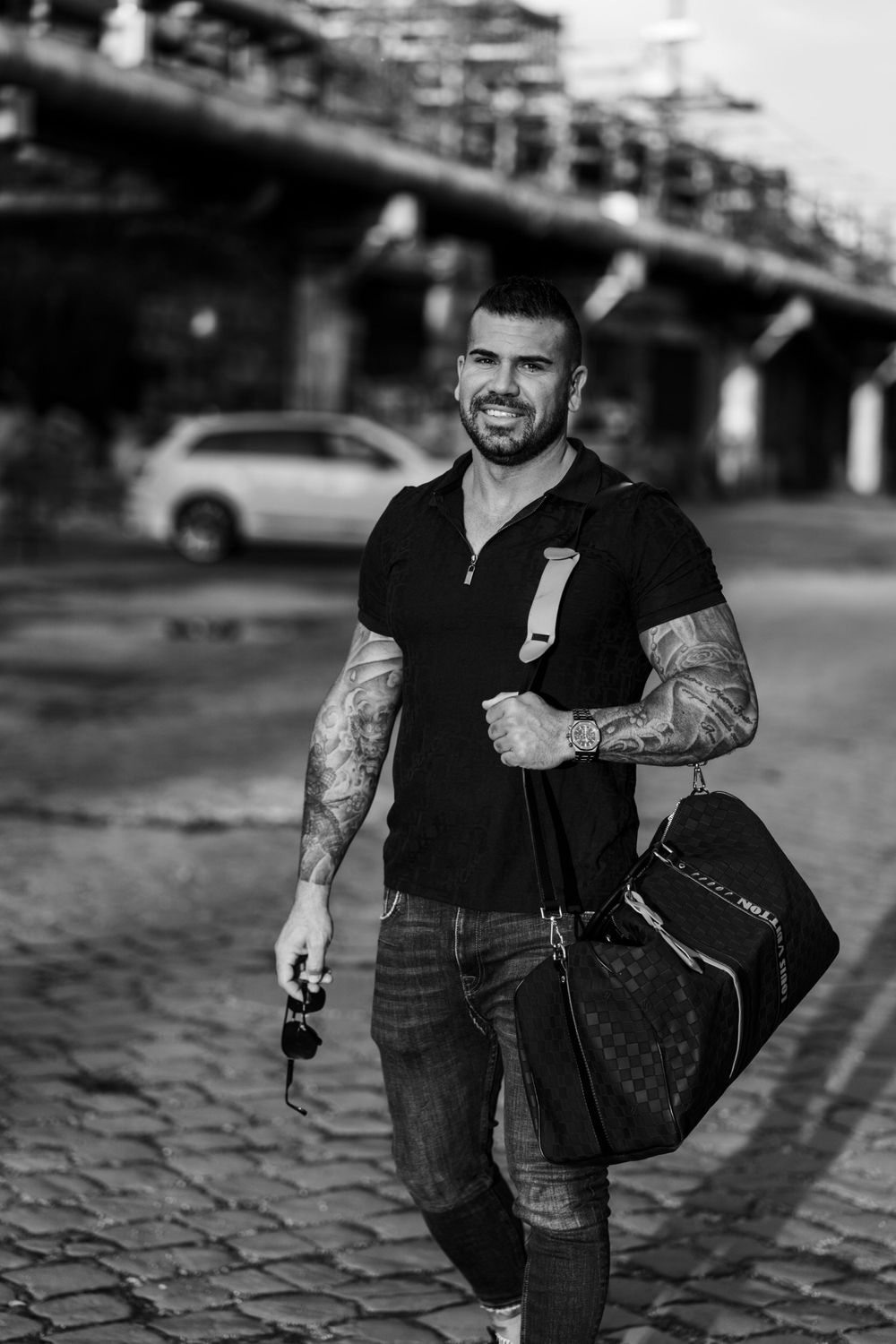Person in black polo shirt carrying designer duffle bag on cobblestone street with urban backdrop.