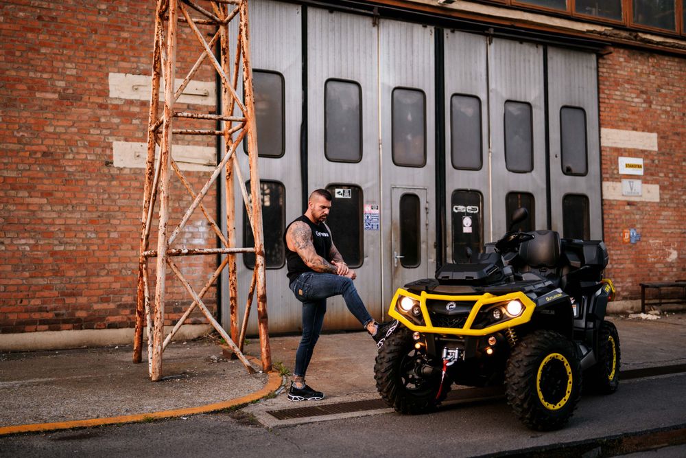 Athletic figure poses next to a yellow and black ATV against an industrial brick building background.