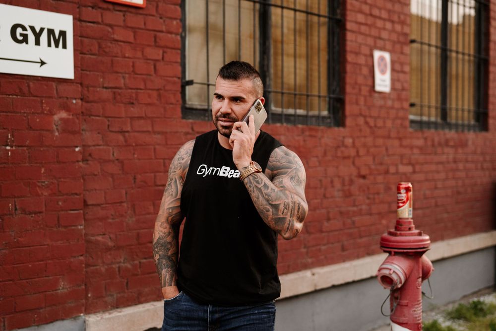 Athletic figure in black tank top stands near red brick wall with gym sign.