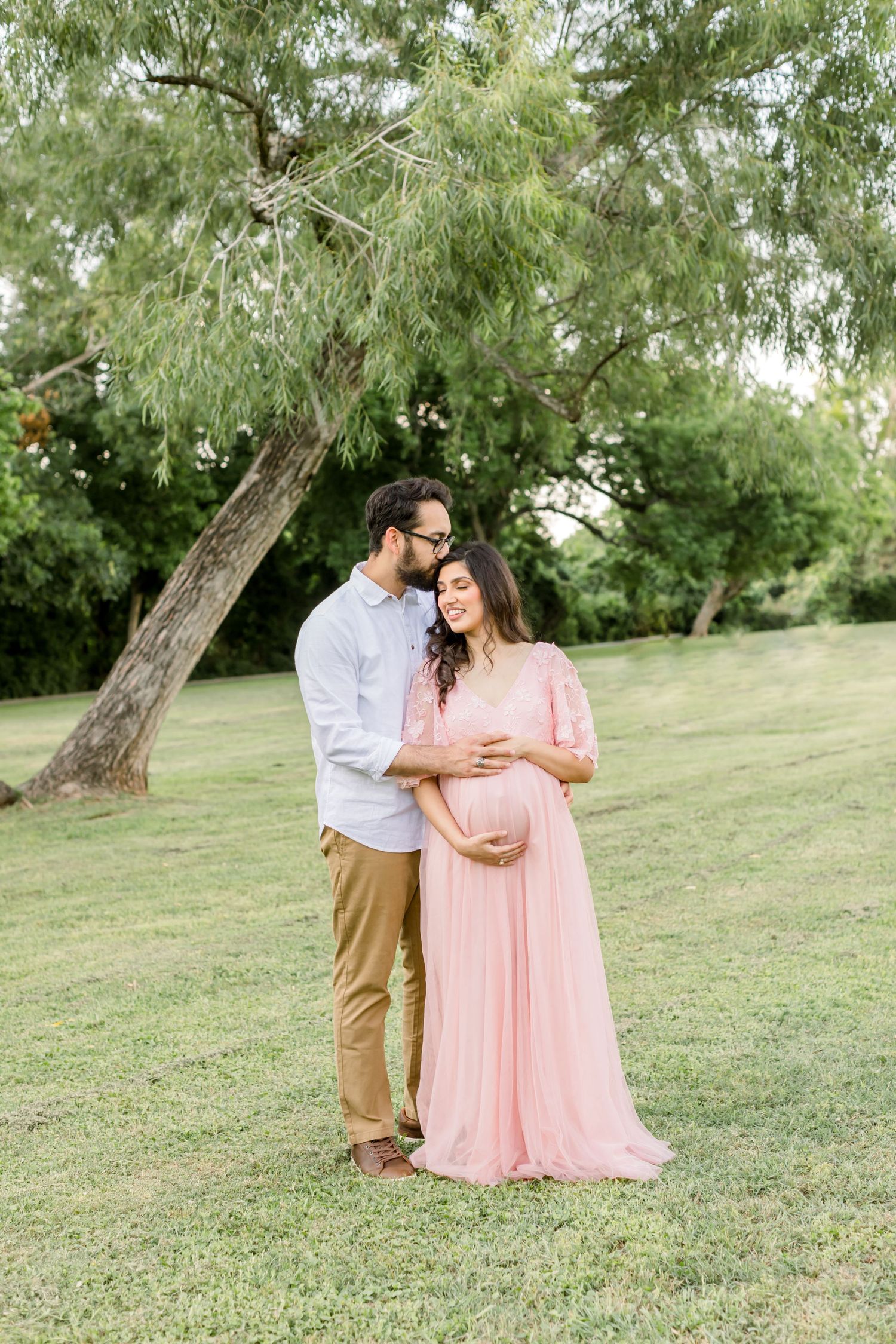 A maternity photo session outdoors featuring a couple embracing under a willow tree on a grassy lawn.
