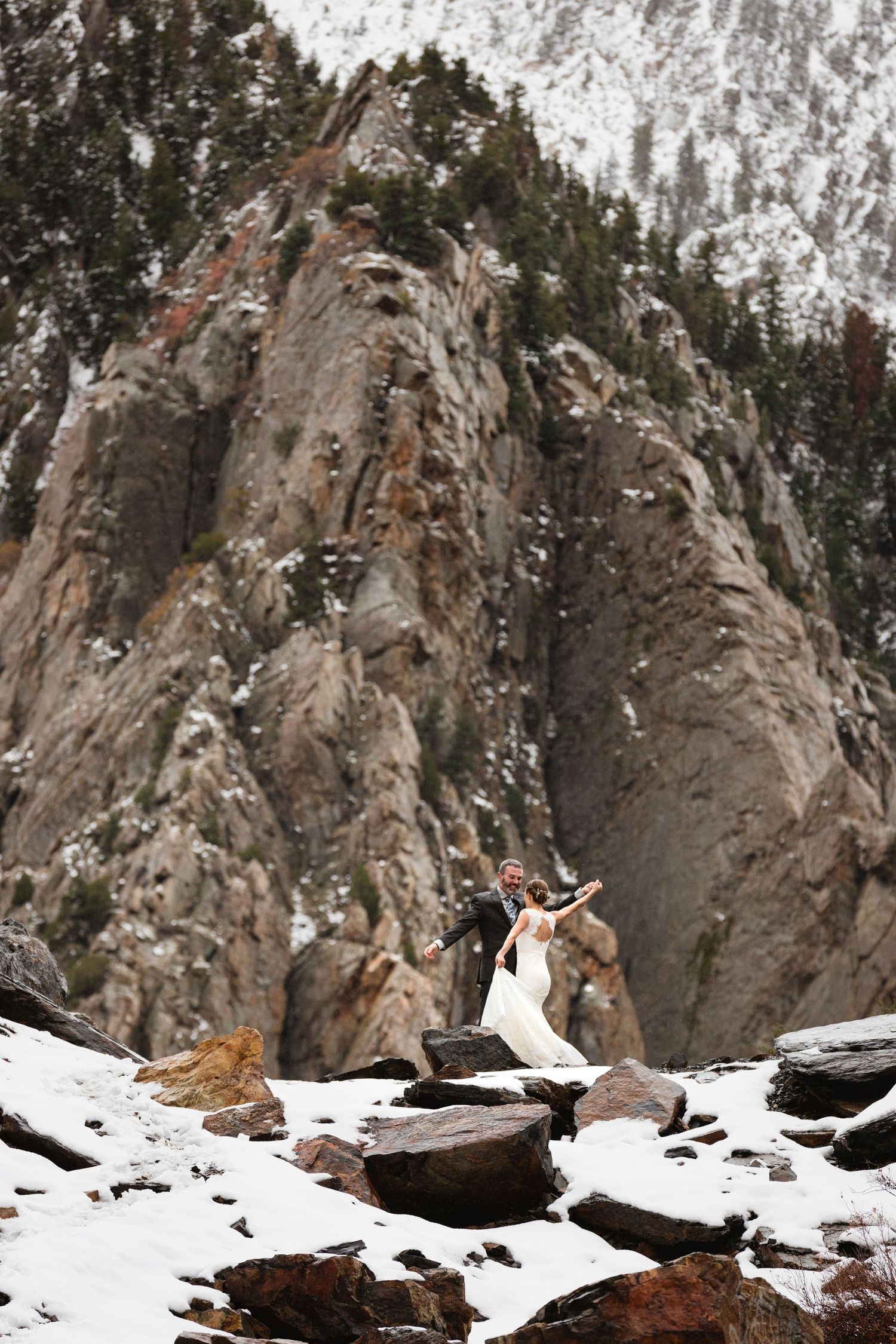 Couple dancing on snowy rocks with dramatic mountain cliff face in winter.
