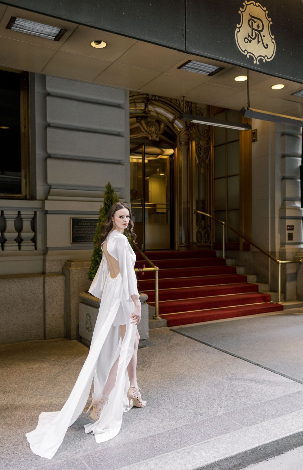 A bride in a flowing white dress stands on red-carpeted steps outside an elegant building entrance.