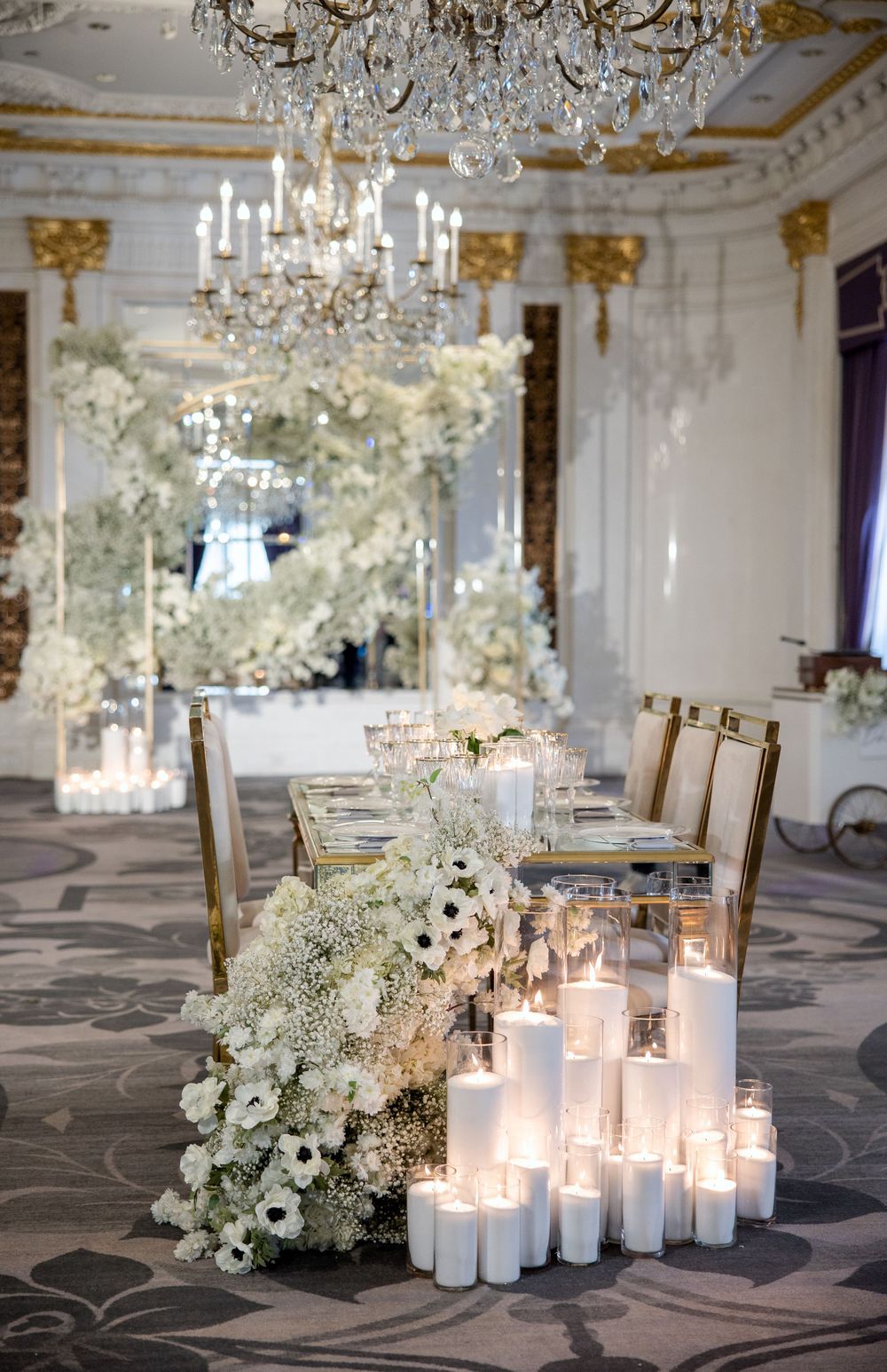 Opulent ballroom wedding decor with crystal chandeliers, white floral arrangements, and glowing candles.