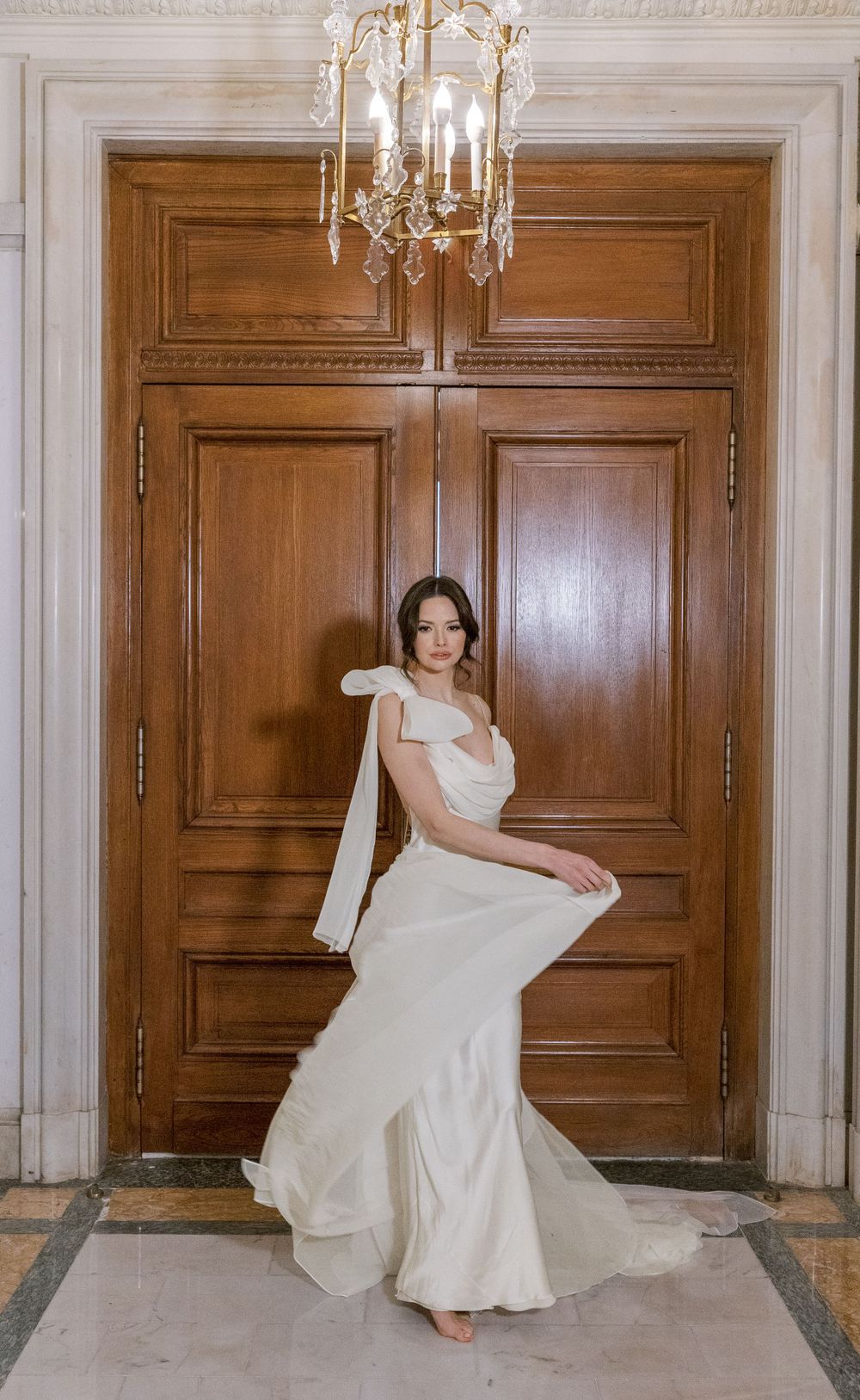 Bride in elegant white gown poses against wooden doors beneath crystal chandelier in classic setting.