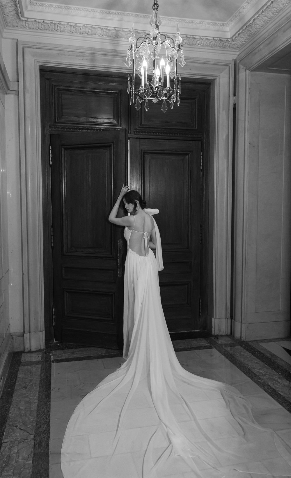 Black and white photo capturing dramatic train of backless wedding gown under vintage chandelier.