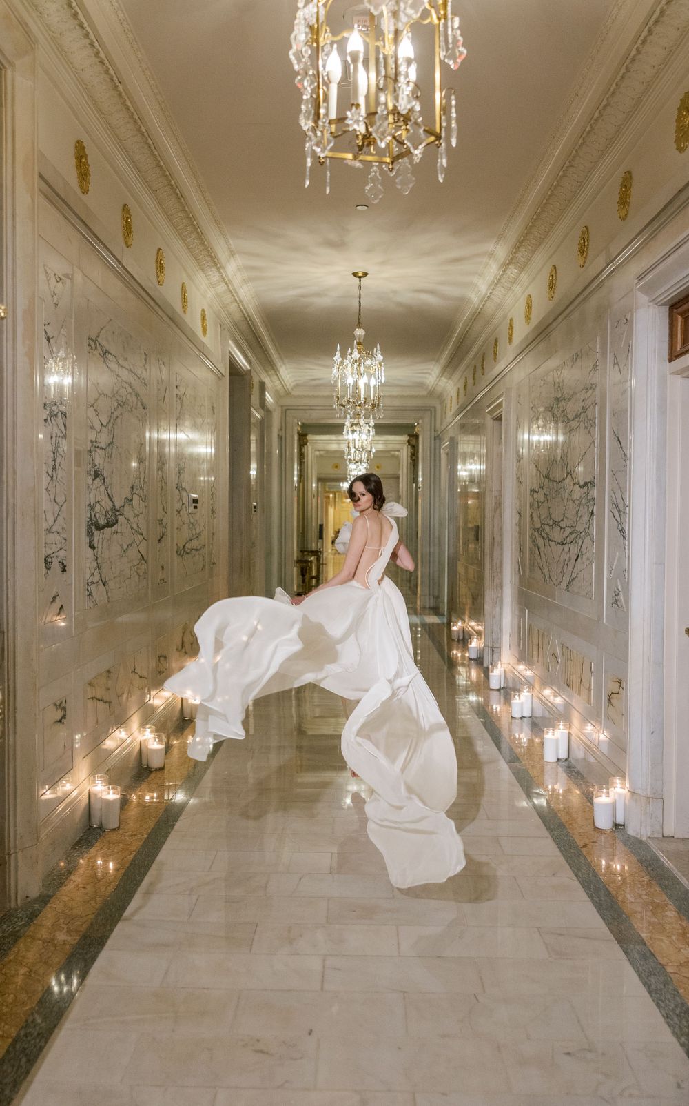 Dramatic bridal dress motion in marble hallway lined with candles and crystal lighting.