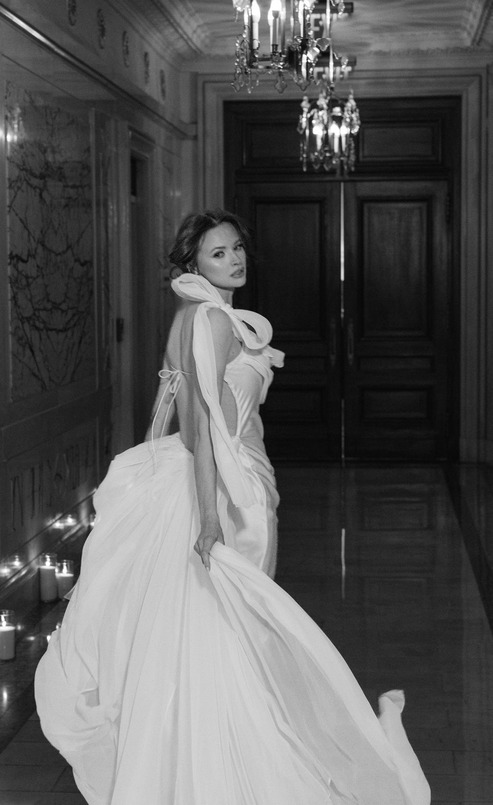 Dramatic black and white photo of a bride in backless gown posing in ornate hallway.