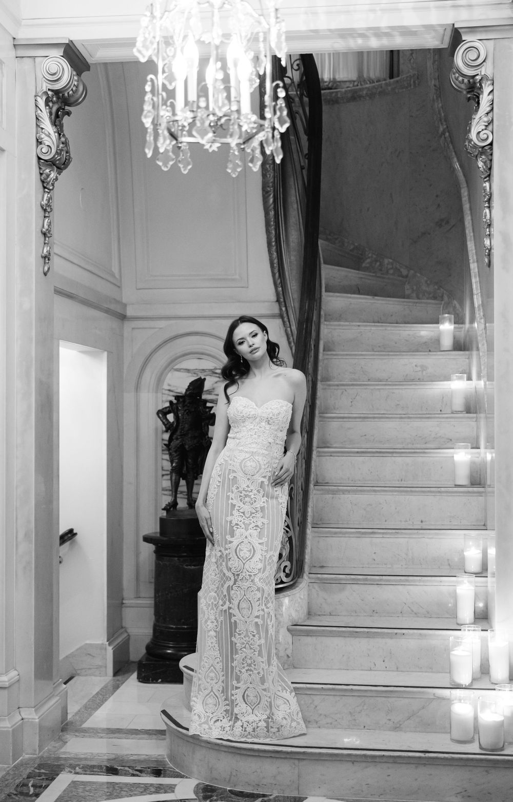 Elegant black and white photo showing a lace wedding gown on a grand staircase with crystal chandelier.