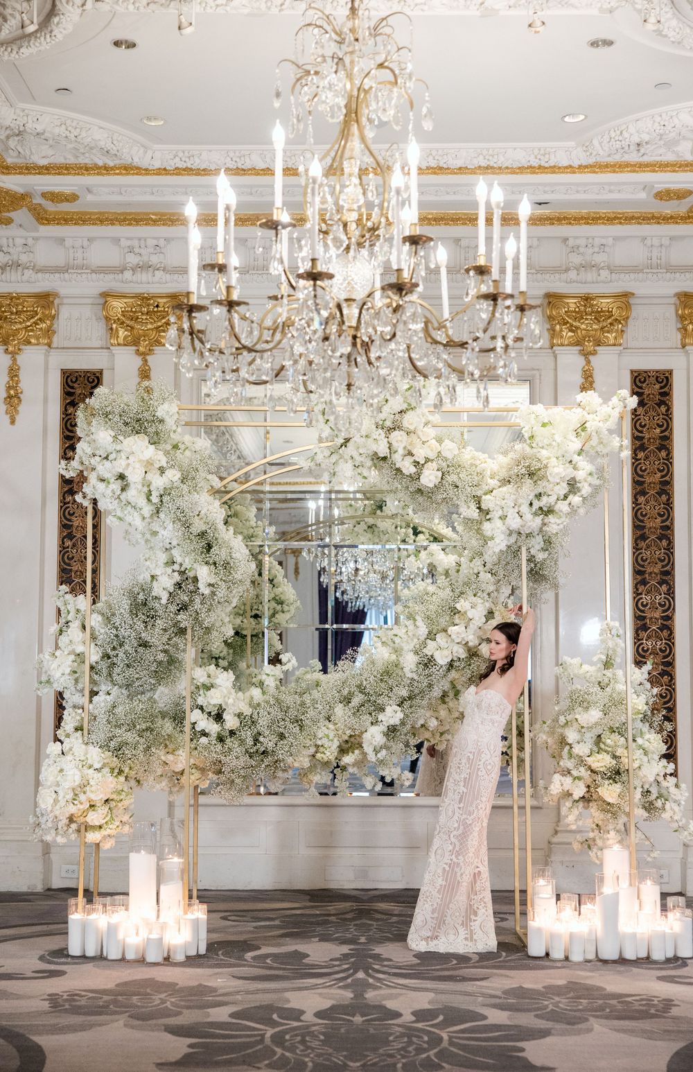 Stunning white floral arch with candlelit base under an ornate crystal chandelier in an elegant ballroom.